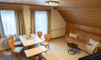 A cozy living space with wooden walls and a table surrounded by chairs. A comfortable couch and a side table complete the relaxed atmosphere.