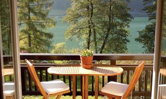 A cozy balcony with a round table and two chairs. In the background, there are trees and a tranquil lake.