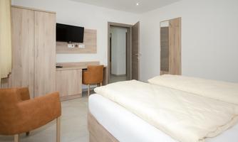 A modern hotel room with a double bed, a desk, and a television. The decor is bright and minimalist.