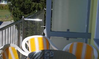 A balcony with two white chairs and a small round table. The chairs have yellow and white stripes, and in the background, trees can be seen.