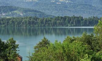 A tranquil landscape with a clear lake and green trees. In the background, gentle hills are visible.