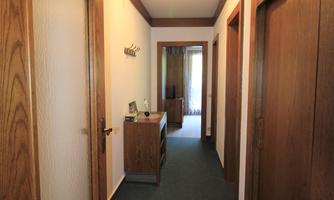 A narrow corridor with wooden doors and carpet flooring. At the end of the corridor is a small table and a window.
