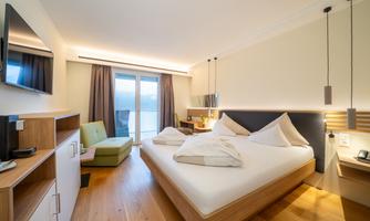 A modern hotel room with a large bed and green seating area. Large windows provide plenty of daylight and a pleasant atmosphere.