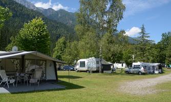 A picturesque campsite surrounded by mountains and trees. The site features motorhomes and a rented accommodation on a green meadow.