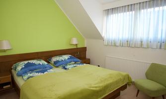 A cozy bedroom with a double bed and green walls. The room has large windows and a modern chair.