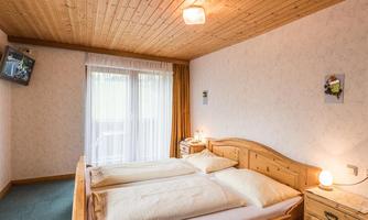 A cozy bedroom with two beds, wood decor, and a large window. The walls are bright, and a TV is mounted on the wall.