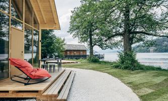 A modern holiday home by the water, surrounded by trees. A cozy terrace with red lounge furniture invites you to relax.