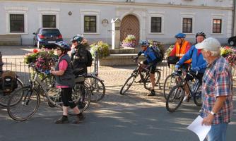 A group of cyclists in athletic clothing is ready to start. In the background, blooming plants and a historic building can be seen.