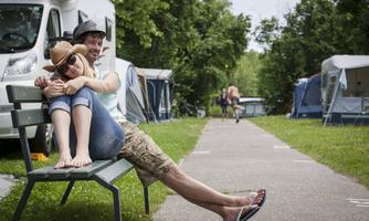 A young couple is sitting on a bench at a campsite. In the background, there are RVs and tents, surrounded by a lot of greenery.