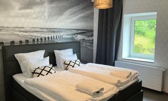 A cozy bedroom with a double bed and decorative pillows. A picture of a coastal landscape hangs on the wall, and a window lets in daylight.