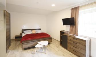 A modern bedroom with a double bed and a red bedspread. Next to the bed is a TV and a small table.