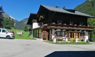 A traditional mountain house with blooming balcony boxes and a picturesque landscape in the background. Surrounded by mountains and green grass, it exudes rural charm.