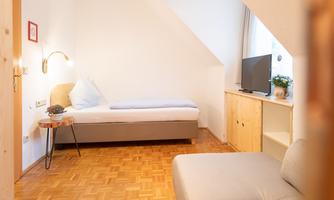 A cozy room with a single bed and a sofa. There is a television and a small wooden chest of drawers.