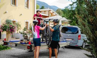 Three people are loading bicycles onto a trailer. In the background, a building and trees can be seen.