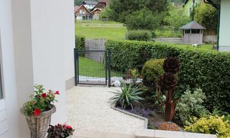 A well-maintained garden with a fence and colorful plants. In the background, some houses and a lot of greenery can be seen.
