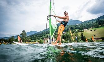 A windsurfer glides across the water, surrounded by green hills and a cloudy sky. In the background, more surfers and a beautiful landscape can be seen.