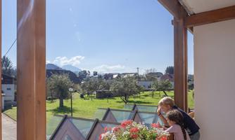 Two children curiously look from a balcony onto a green meadow with trees and houses. The sun shines brightly in the blue sky.