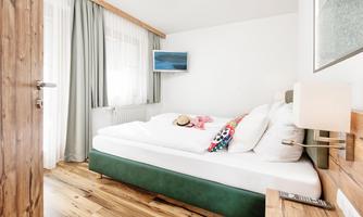 A cozy room with a large bed and wooden flooring. A television hangs on the wall and the windows are decorated with curtains.