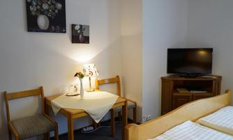 A cozy room with a bed, table, and chairs. Pictures hang on the wall, and a television stands on a wooden cabinet.