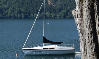 A sailboat lies peacefully on a clear lake, surrounded by green trees. In the background, gentle mountains can be seen.