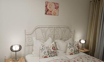 A cozy bedroom with a white bed and colorful pillows. A picture of roses hangs on the wall, and the nightstands are equipped with modern lamps.