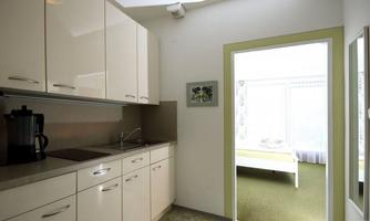 A modern kitchen with white cabinets and a countertop. In the background, a bright room with a bed can be seen.