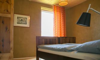 A cozy bedroom with a double bed, a floor lamp, and a window with colorful curtains. An artwork hangs on the wall, and the walls are in warm tones.