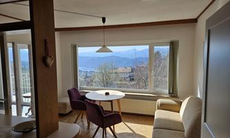 A bright and inviting living room with a round table and modern chairs. The view through the window showcases a picturesque landscape with mountains and a lake.