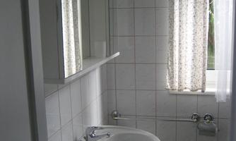 A small, bright bathroom with a sink and a mirror. It has white tiles and a window with a curtain.
