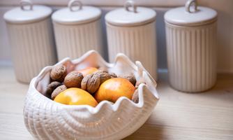 A decorative bowl with various fruits and nuts is sitting on a table. In the background, four identical containers are visible.