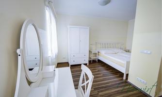 A simple bedroom with a white bed, a desk, and a mirror. The room has light walls and a wooden floor.