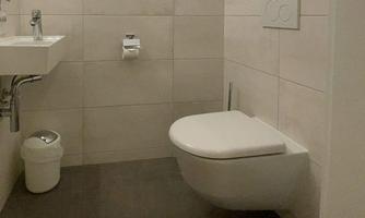 A modern bathroom with a wall-mounted toilet and a small sink. The walls are light tiled and the floor is dark.