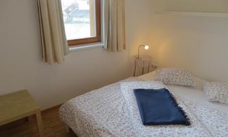 A simple bedroom with a bed and a table. There is a window side with curtains and a bedside lamp.