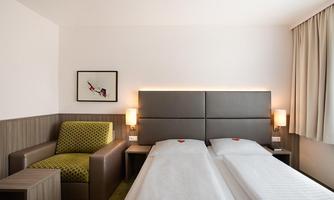A comfortable hotel room with two single beds and a cozy armchair. The wall is bright and modernly designed, with soft lighting.