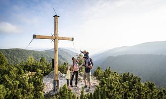 A family is standing on a summit next to a wooden cross. In the background, green mountains and a sunny, cloudless sky can be seen.