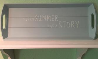 A gray tray with the inscription "Every summer has a story." It is placed on a white shelf.