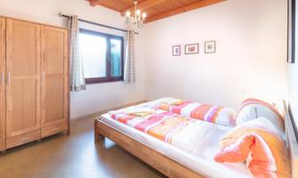 A cozy bedroom with a double bed and colorful bedspreads. A wooden chest of drawers and a window with curtains create a bright atmosphere.