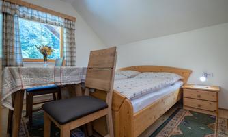 A cozy room with a single bed, a table, and a chair. Daylight comes through the window and there is a beautiful view of the surroundings.