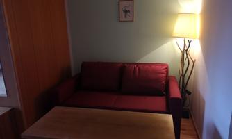 A cozy living room with a red sofa and a wooden table. There is a lamp on the wall that provides pleasant light.