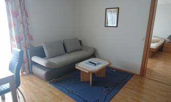 A bright living room with a gray sofa and a table. In the background, a bedroom can be seen.