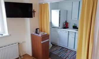 A small kitchen with modern appliances and a colorful carpet. In front of the kitchen, there is a table with a coffee machine.
