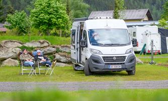 A motorhome is parked on a green campsite. Two people are sitting relaxed in folding chairs.