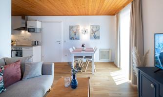 A modern apartment with an open kitchen and a cozy living area. The dining table is bright and inviting, surrounded by windows for plenty of natural light.