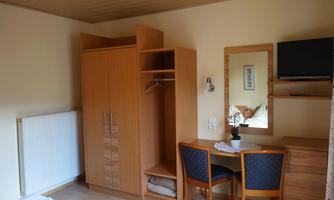 A simple bedroom with wooden furniture. There is a wardrobe, a table with chairs, and a television on the wall.