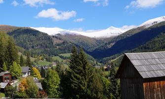 A picturesque mountain landscape with snow-capped peaks and green forests. In the foreground, traditional wooden houses and a bright blue sky can be seen.