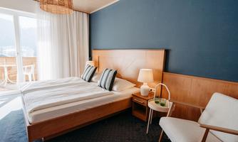 A modern bedroom with a double bed and wooden details. The room has large windows with curtains and a cozy sitting area.