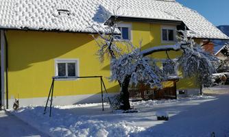 A yellow house in the snow with a snow-covered tree and a swing in the foreground. The sky is clear and blue.