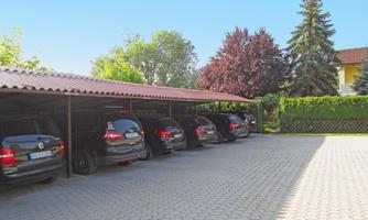 A row of black cars is parked in a covered parking lot. In the background, trees and a building are visible.