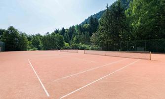 A tennis court with a red clay surface, surrounded by trees and green hills. The sky is clear and the atmosphere is calm.