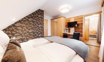 A cozy bedroom with a large bed and wooden paneling. In the background, there is a TV unit and a desk.